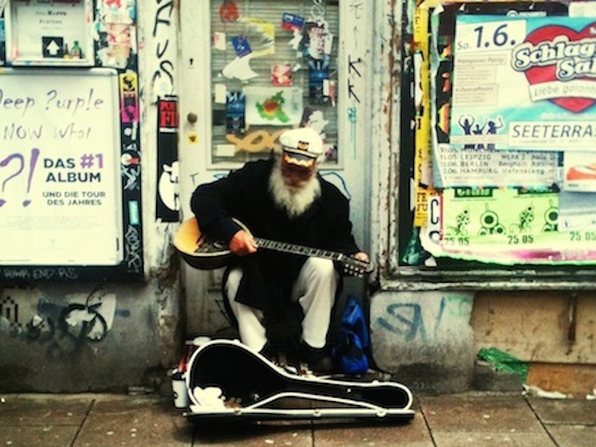 Ein Straßenmusiker mit Saiteninstrument, weißem Rauschebart und Kapitänsmütze musiziert im Hamburger Schanzenviertel vor einer plakatierten Wand