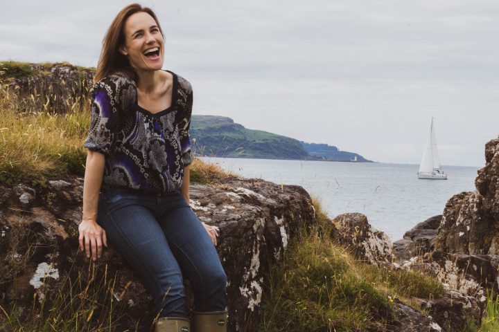Whisky-Produzentin Annabel Thomas sitzt lachend auf einem Felsen vor der schottischen Küste