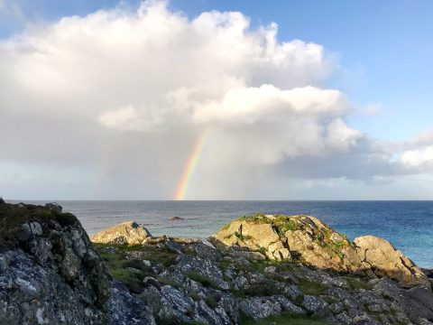 Tiree: Ein Regenbogen kommt unter flauschigen Wolken hervor und landet im blauen Meer